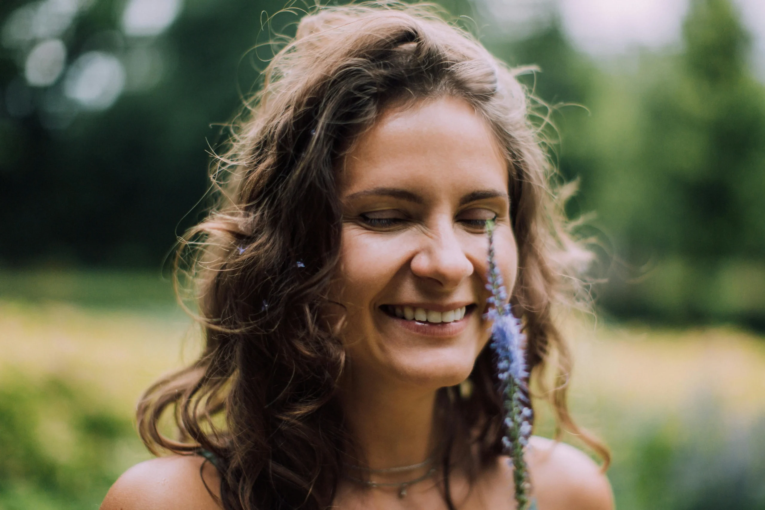 young woman in nature with eyes closed smiling as she smells a wildflower